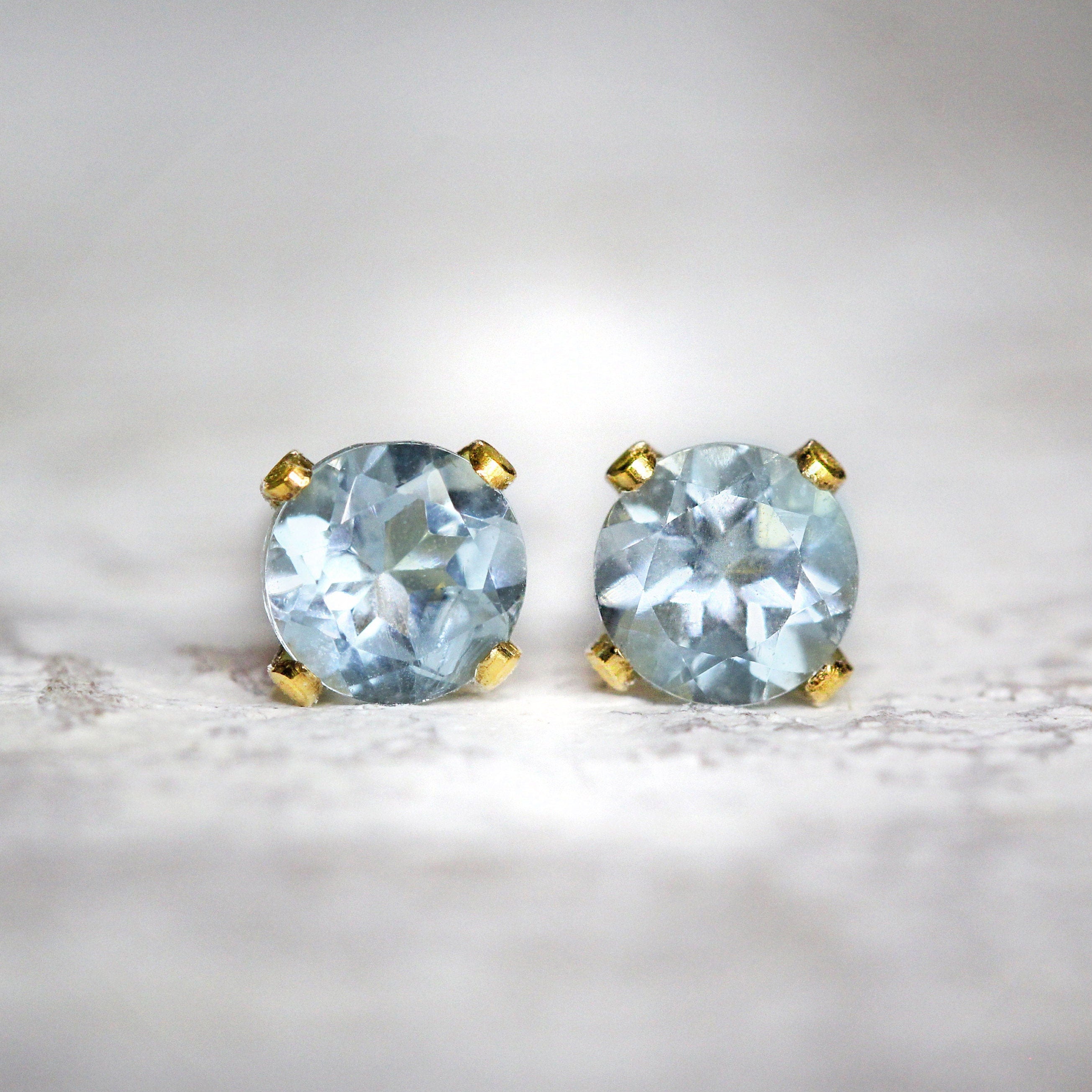 A pair of matching gemstone stud earrings in gold settings. The gemstone is round, faceted, sparkling and pale blue in colour. The stones are set into 14k gold fill ear posts with a 4 prong setting. They come with matching butterfly backs.
