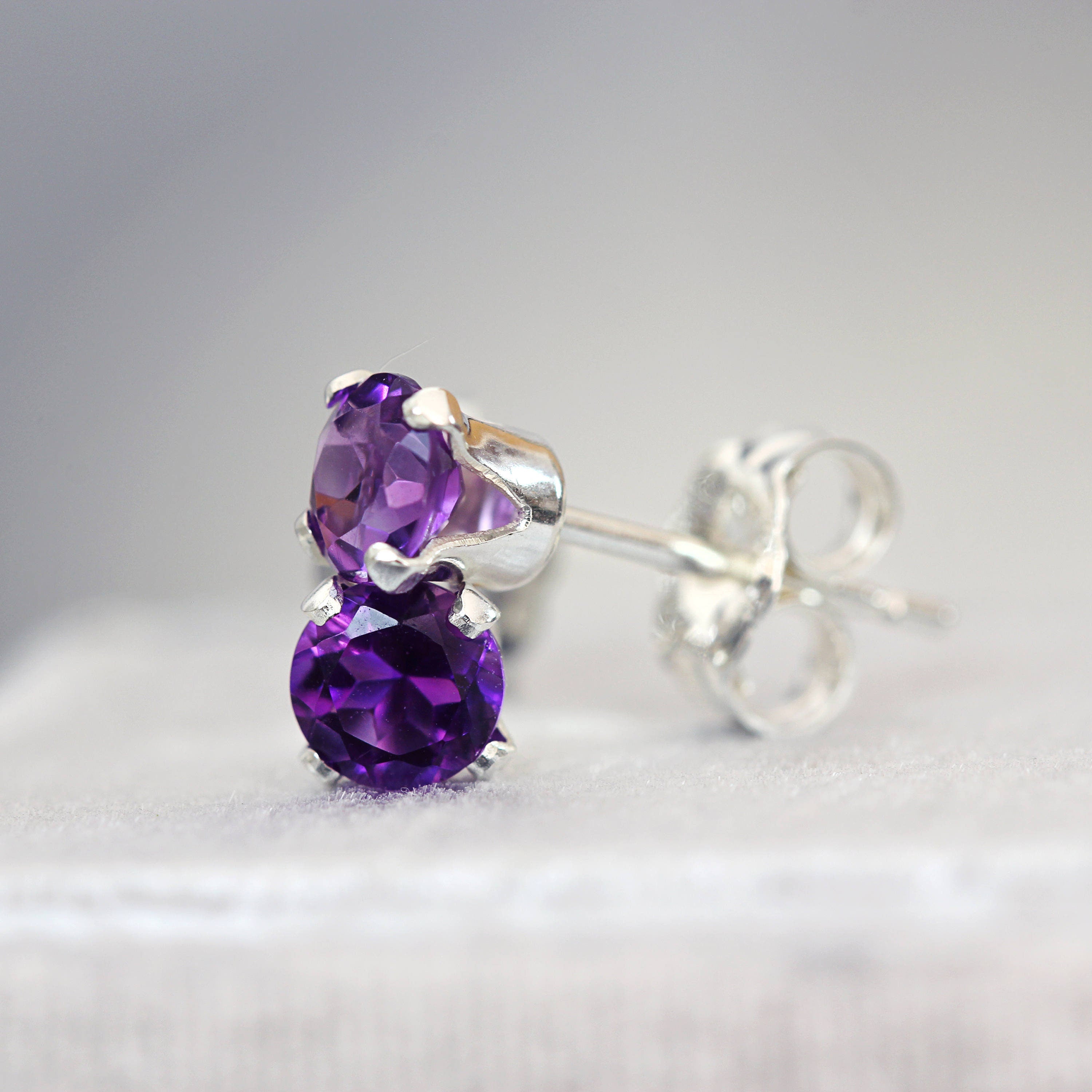 A pair of dainty silver stud earrings featuring round deep purple amethyst gemstones in a prong setting. Round in shape, with ear post fitting and matching butterfly backs. The earrings are simple, sophisticated and minimal and come in various sizes.
