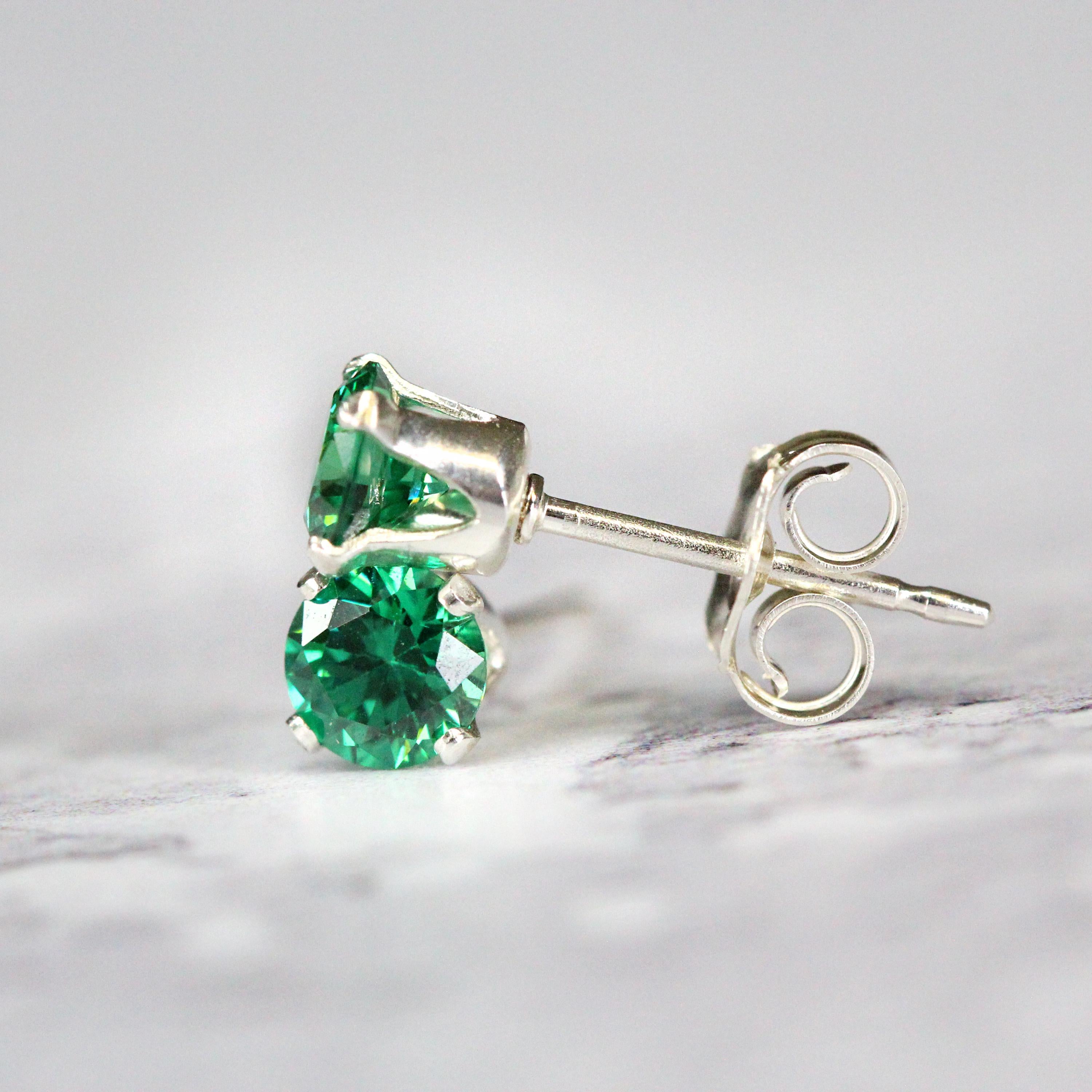 A pair of emerald green cubic zirconia stud earrings. The cz gemstones are sparkling and catch the light. The stones are a brilliant round cut with lots of facets, and set into sterling silver four prong settings. Simple, minimal, round stud earrings.