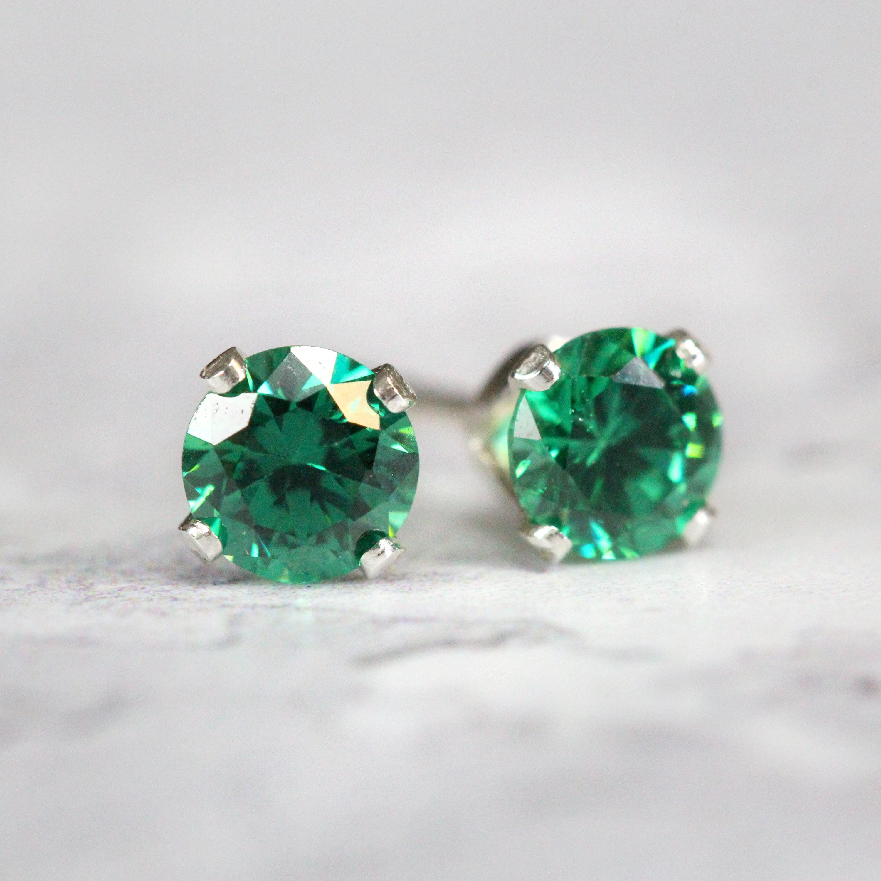A pair of deep teal green cubic zirconia stud earrings. The cz stone is round in shape with a faceted cut. The sterling silver ear post setting has four prongs holding the gemstone in place, and butterfly push backs to hold the stud securely on the ear.