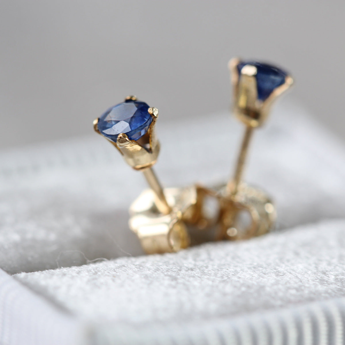 A pair of blue sapphire stud earrings with gold-colored metal posts and butterfly backs, displayed on a white surface.